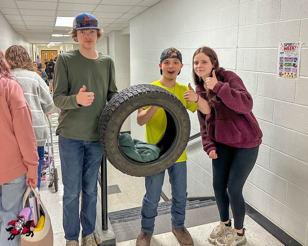 Three teens, one wearing a cap, pose behind a large tire. A woman with a bag is in the foreground.