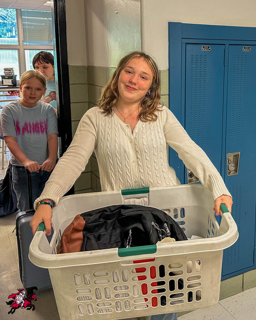 A young girl carries a basket of laundry in a hallway. Two children stand behind her, one holding a suitcase. Lockers are on the right side.