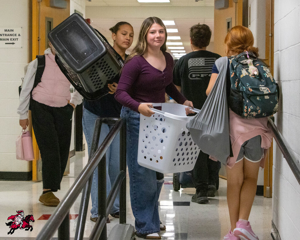 Students in a hallway, carrying a laundry basket, a cat carrier, and backpacks. Hallway has doors and handrails.