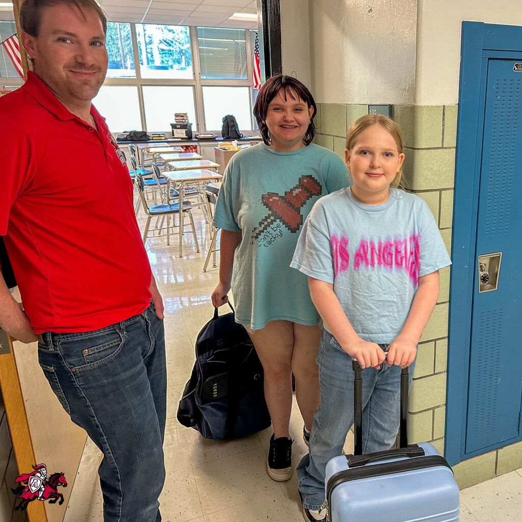 A man and two girls stand in a hallway with lockers. The man wears a red shirt, the girl in front a gray tee.