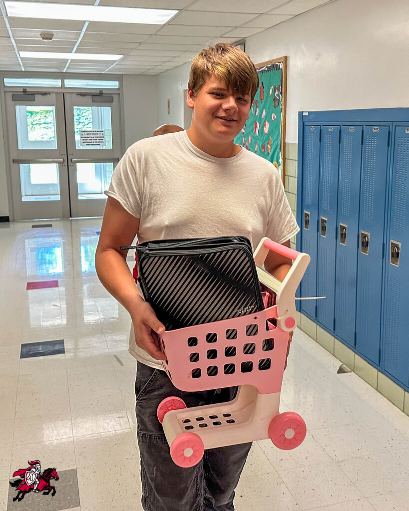 A person holding a pink toy shopping cart filled with items, standing in a hallway with lockers.
