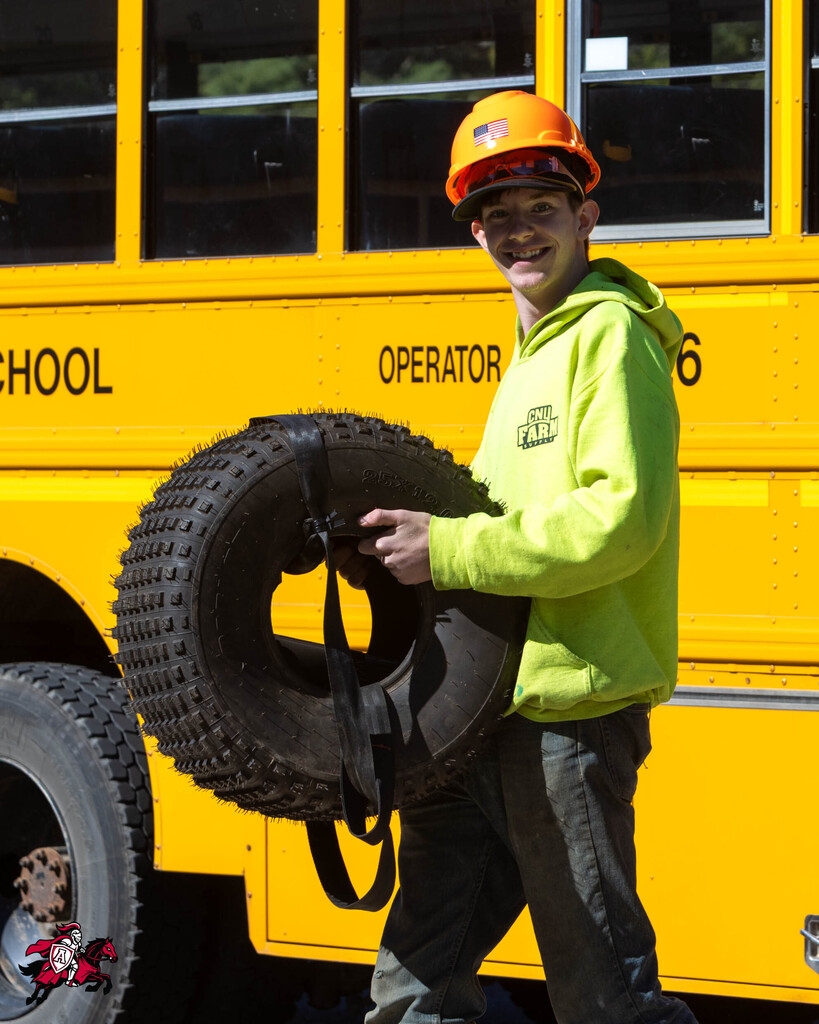 Man in a yellow sweatshirt, orange helmet, and pants carrying a large tire in front of a yellow school bus.