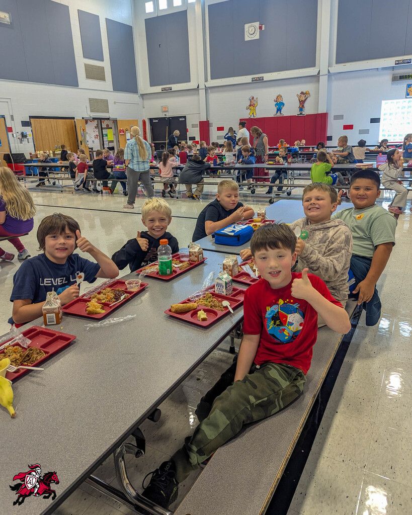 Children sitting at cafeteria tables with trays of food. A boy in a red shirt gives a thumbs-up.