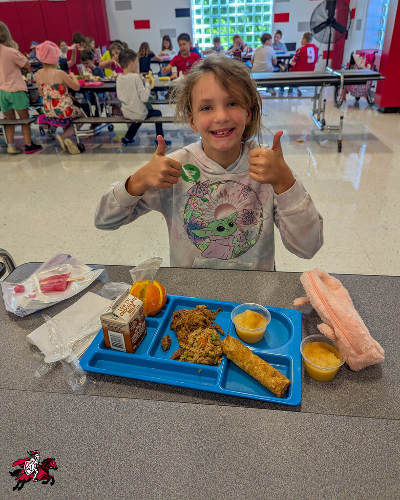 A young girl gives a thumbs-up in a cafeteria. She sits at a table with a blue tray of food.