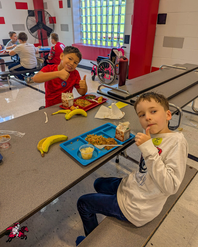 Two young boys seated at a table in a cafeteria with trays of food, one smiling and the other with his hands near his mouth.