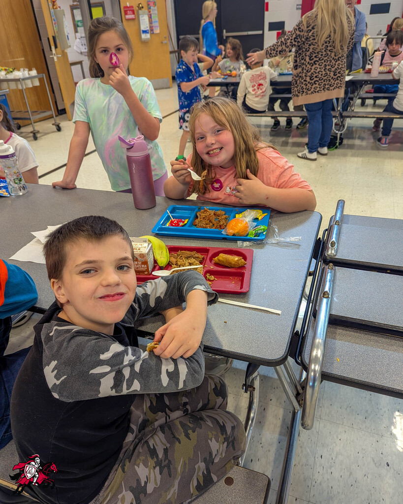 Kids eating at a school lunch table. One child holds a sandwich. Other children stand or sit in the background.