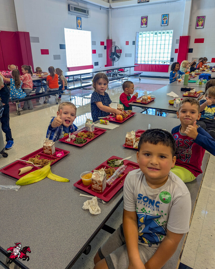 A group of children in a cafeteria with tables, food, and red trays. Bright windows and red walls.