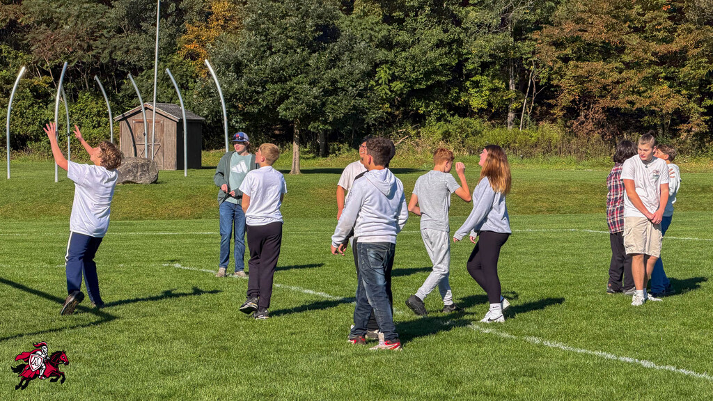 Group of people in casual clothes on a field, some with arms raised, others walking, with trees in the background.