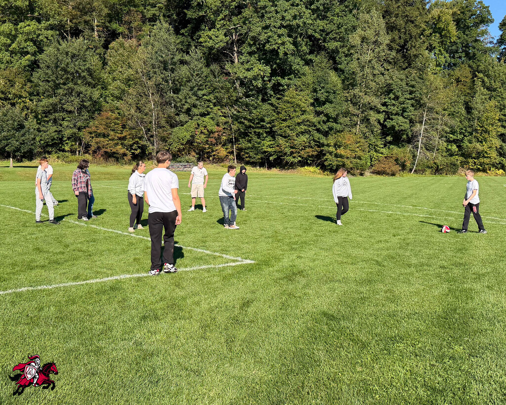People stand on a field, divided by white lines. Trees are in the background. A red and white object lies on the grass.