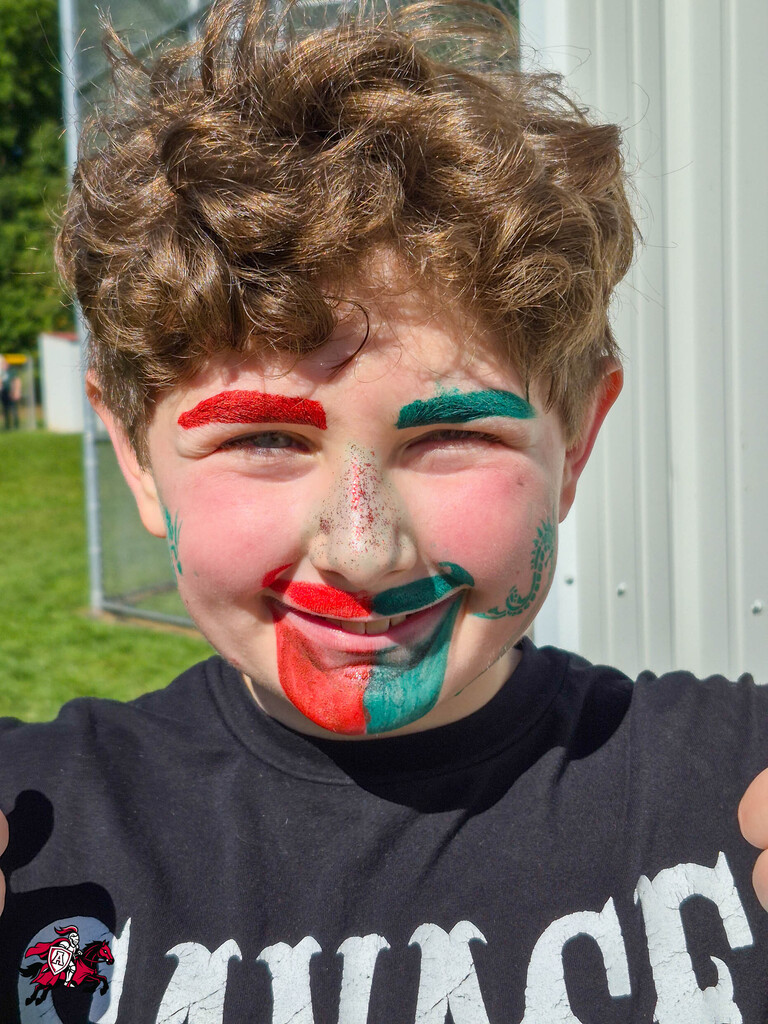 A young boy with curly hair, wearing a black shirt, has a face painted in red, green, and white.