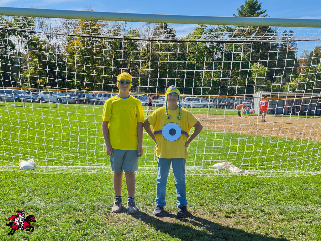 Two people dressed in yellow pose in front of a soccer goal, with a green field, parked cars, and trees in the background.