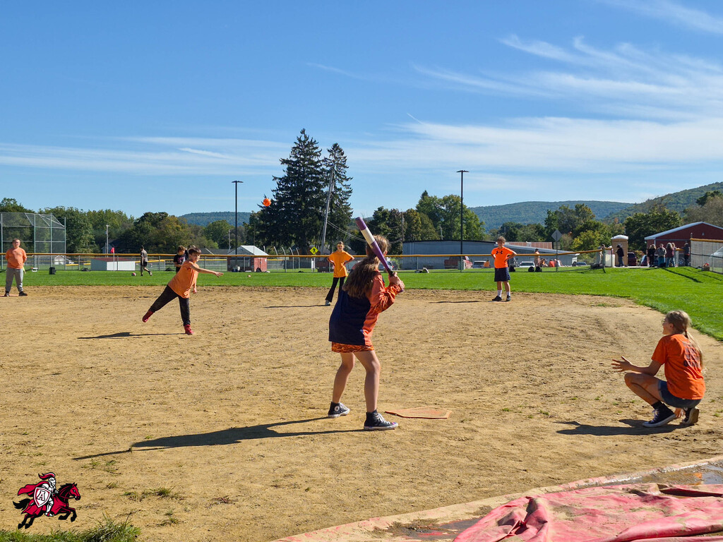 Children playing softball outdoors, on a dirt field with shadows. One holds a bat, another crouches. Background includes trees, grass, and mountains.