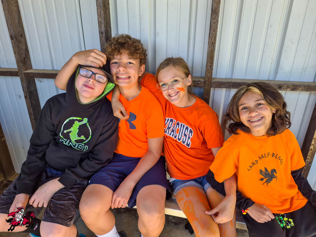 Four children in orange T-shirts pose for a photo in front of a metal wall. One child wears glasses.