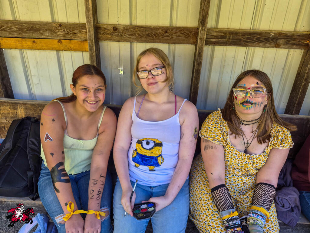 Three women sit on a bench. One has face paint, another wears a Minion shirt, and the third has bracelets and a handbag.