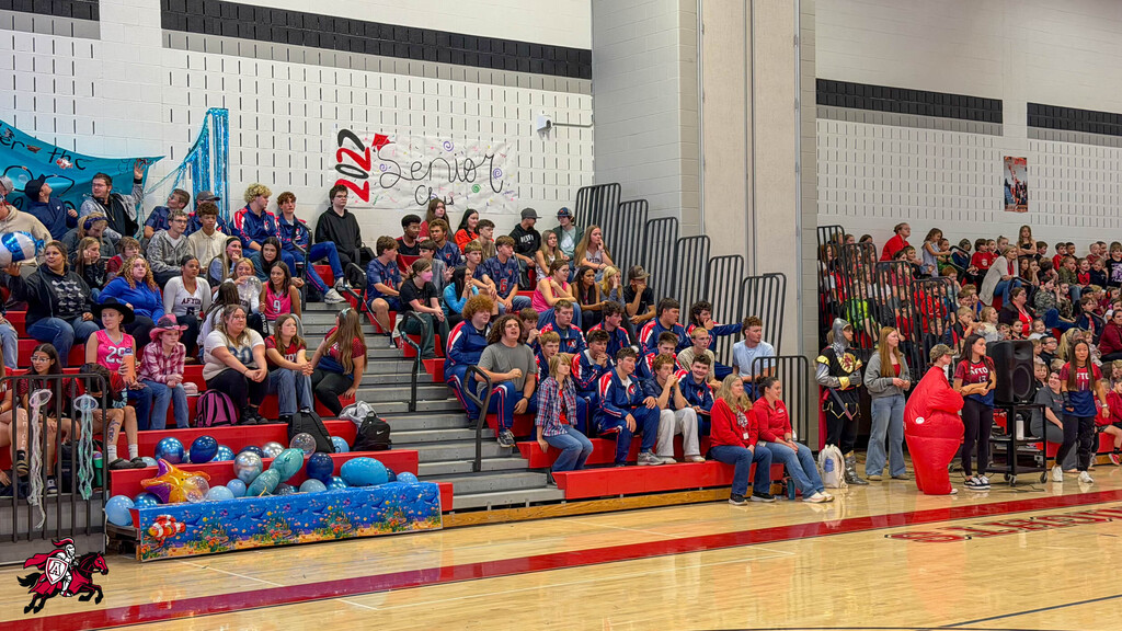Large crowd seated on bleachers in a gym, some wearing red and blue uniforms, with decorations and a mascot present.