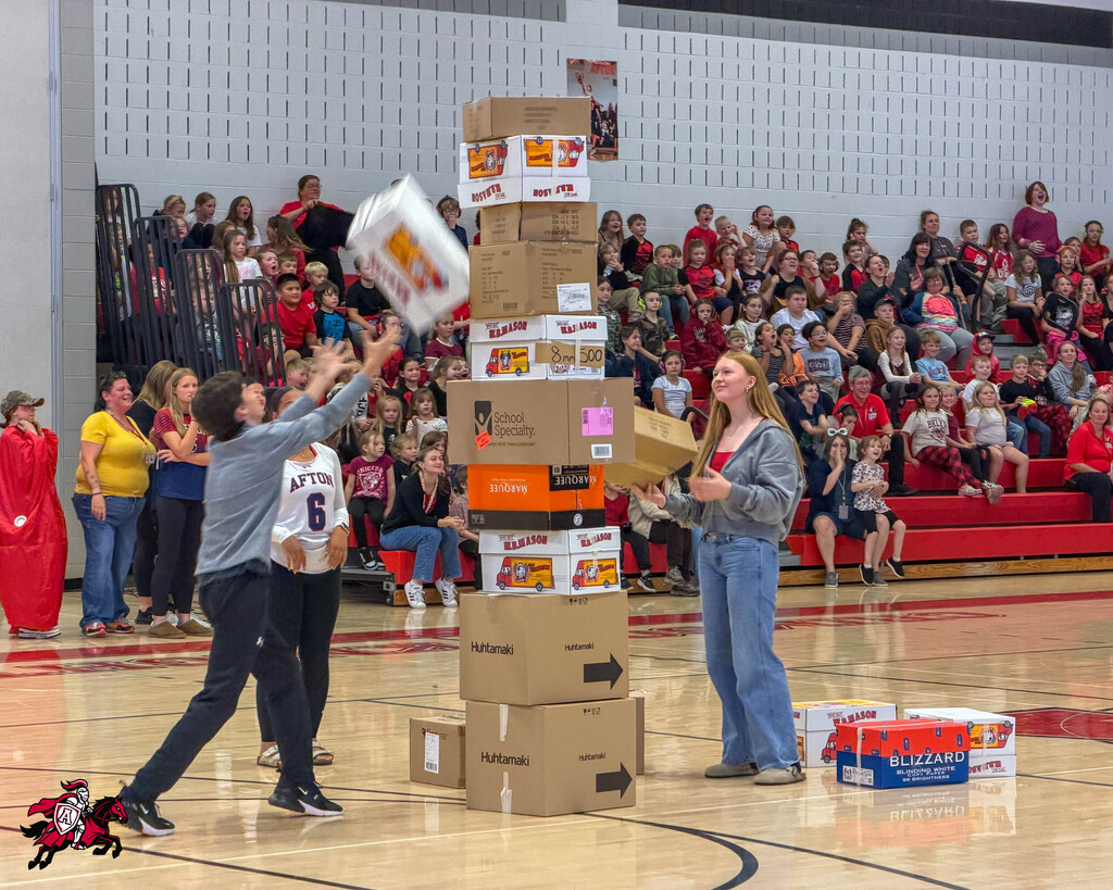 A group of people inside a gymnasium stacking boxes. Audience seated in bleachers watching.