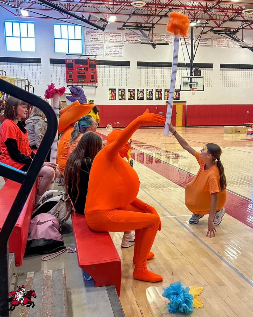 A person in orange holds a pom-pom, raising it high above their head in a gym.