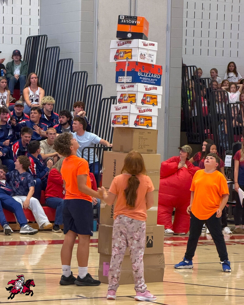 A team of four people in orange shirts stacks boxes on a basketball court as a crowd watches.