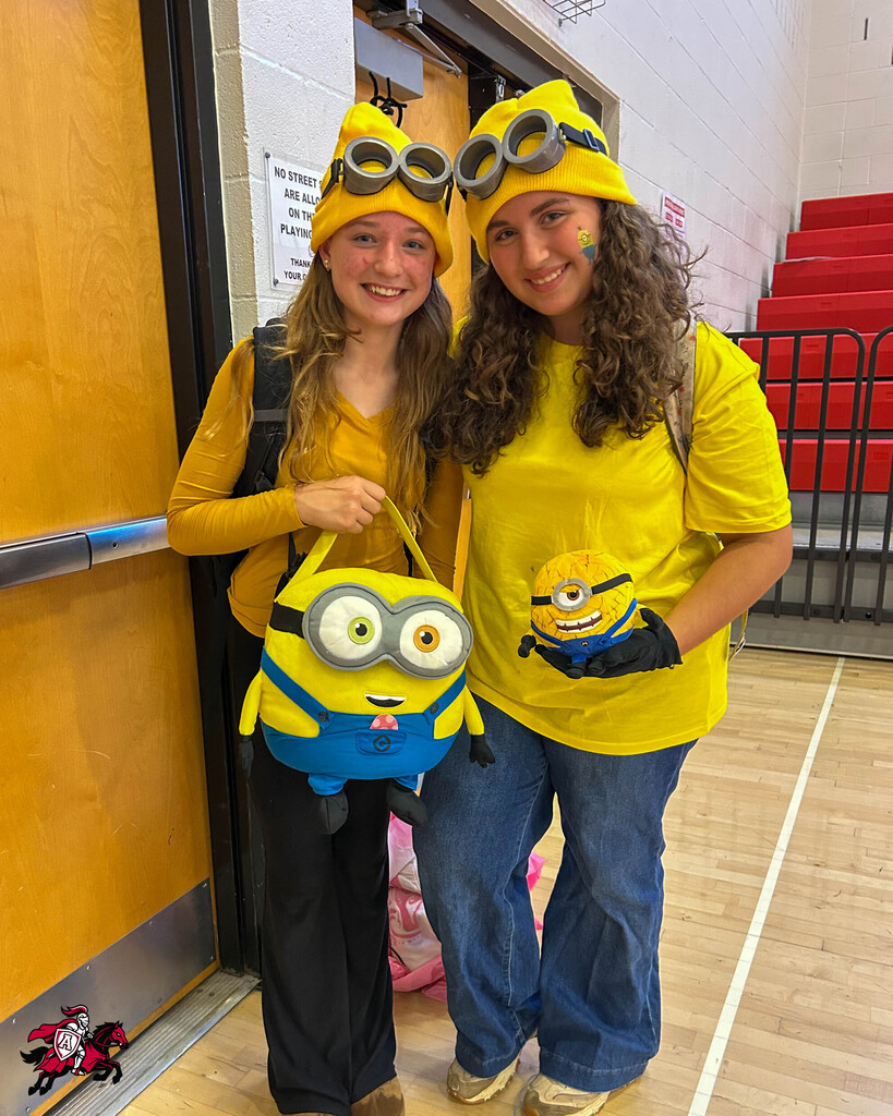 Two women in Minions costumes stand in a gym, one holding a Minion toy. Behind them, a door and a red bleacher are visible.