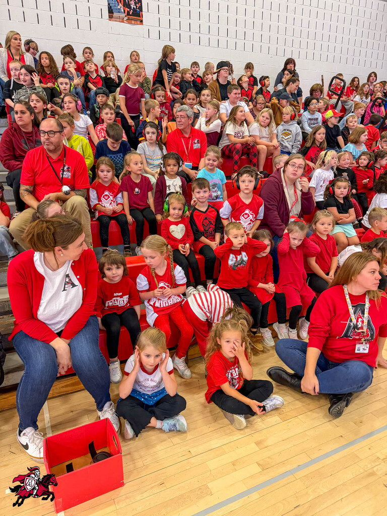 Group of children and adults in red shirts seated on bleachers and floor, possibly at a school event.