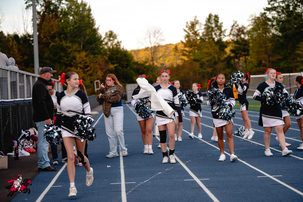 Cheerleaders in uniform, with red bows in their hair, run on a track, with trees in the background.