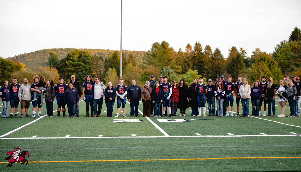 A group stands on a football field, with several people wearing navy blue shirts. A mountain with trees is in the background.