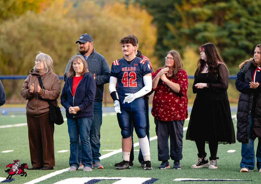 People stand on a field with a football player in uniform, hands clasped, during a pre-game ceremony.