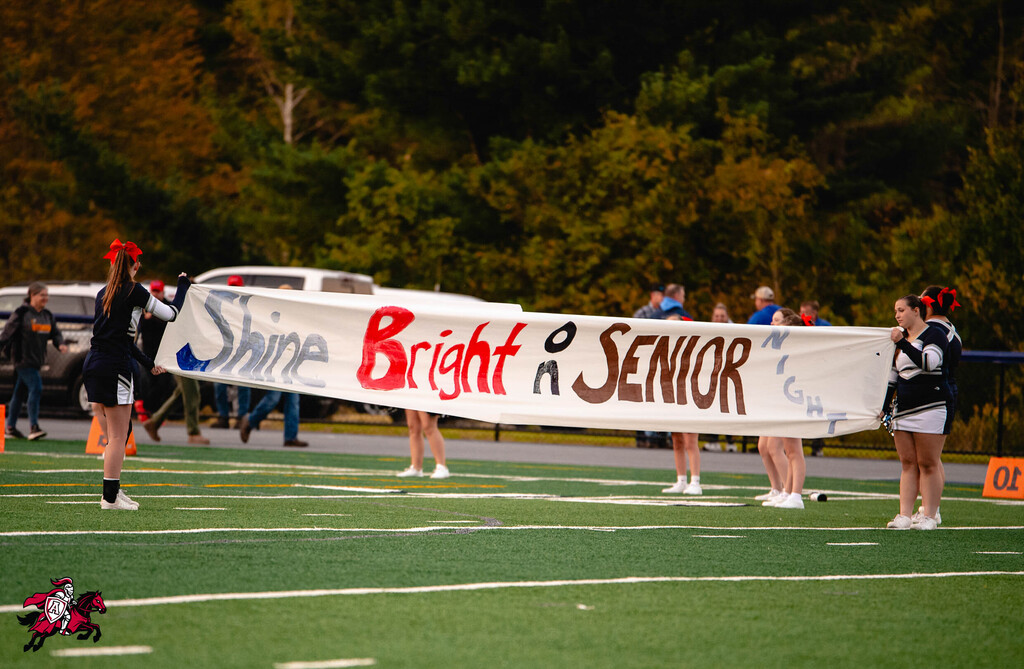 A group holds a banner reading "Shine Bright as Senior" on a football field, surrounded by blurred trees.