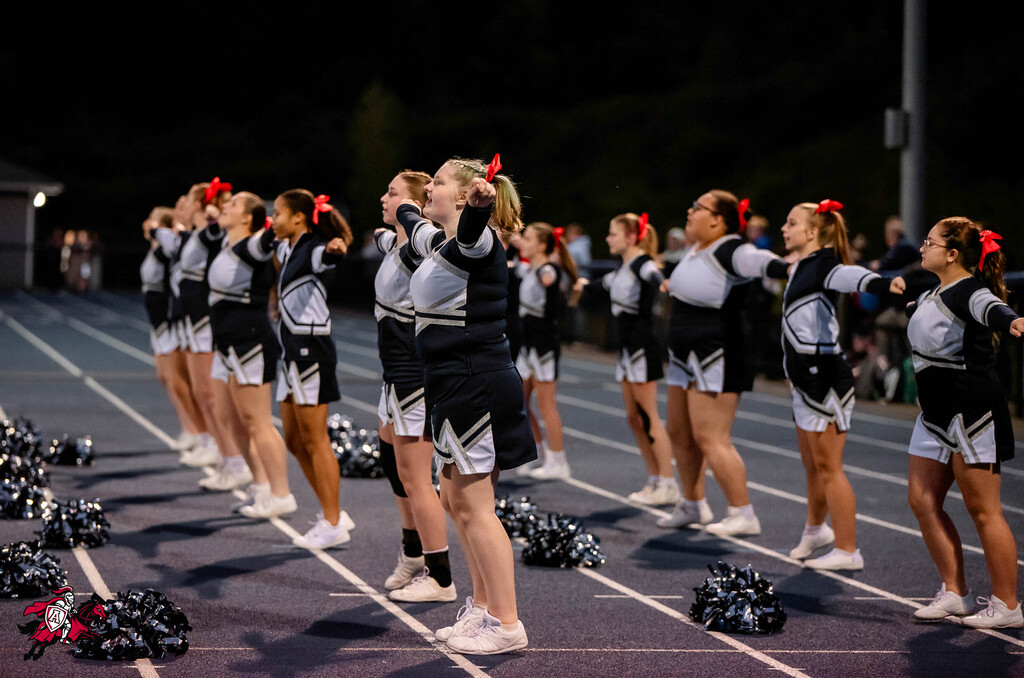 A group of cheerleaders in matching uniforms stands on a track. They are holding pom-poms and looking upward.