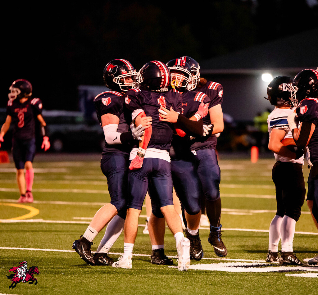 Players in dark uniforms, helmets, and sneakers embrace on a field. Others stand nearby, watching.