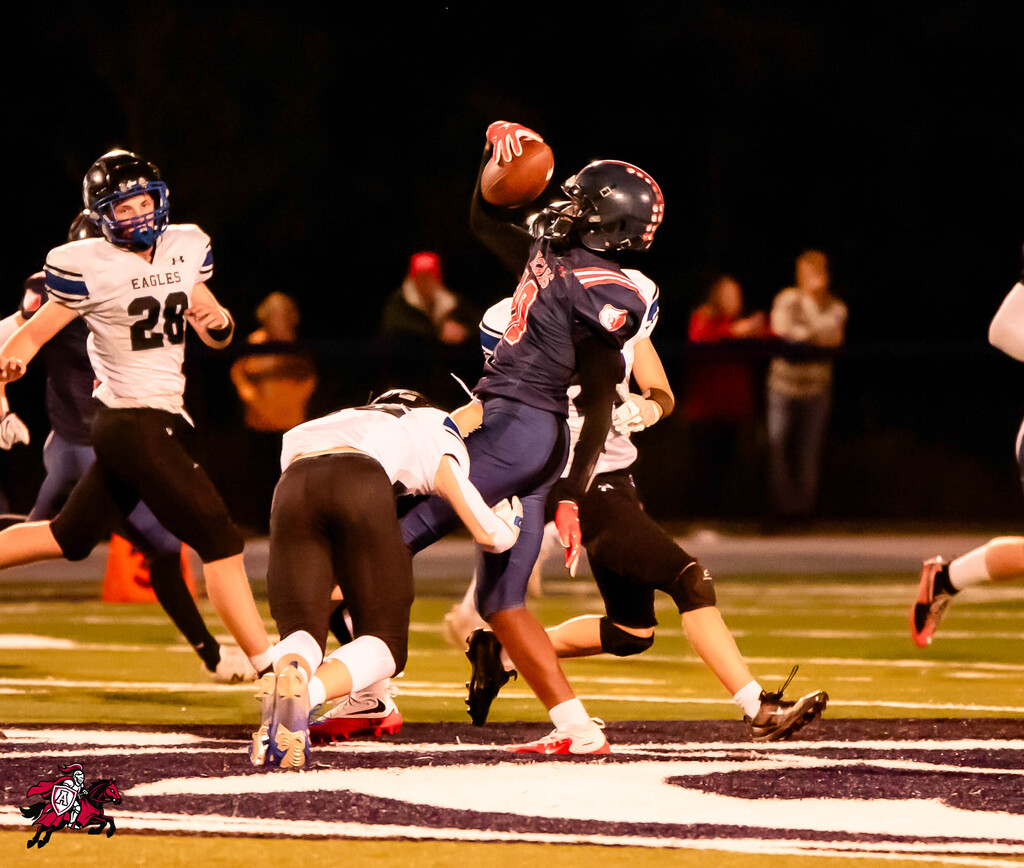 Football players on a field at night; one player in blue prepares to throw a ball. Spectators stand behind the fence.