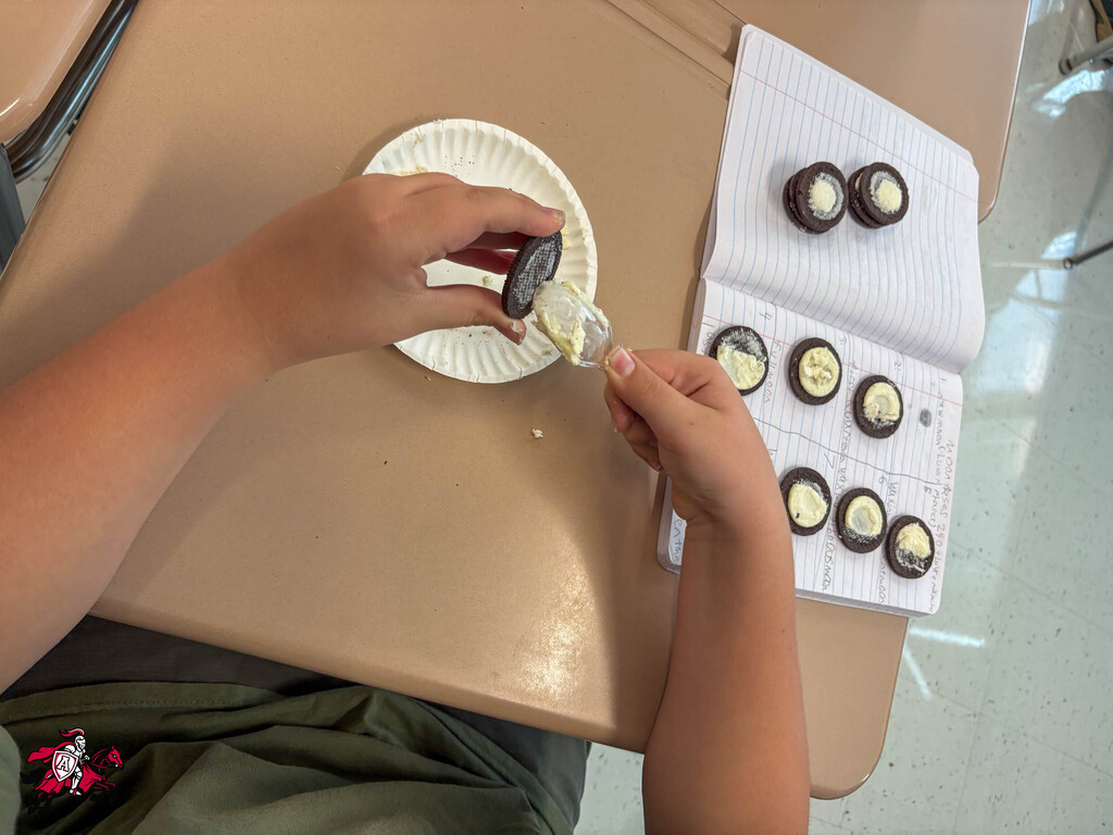 Person at a table, decorating a cookie with icing, using a paper plate, with a book open.