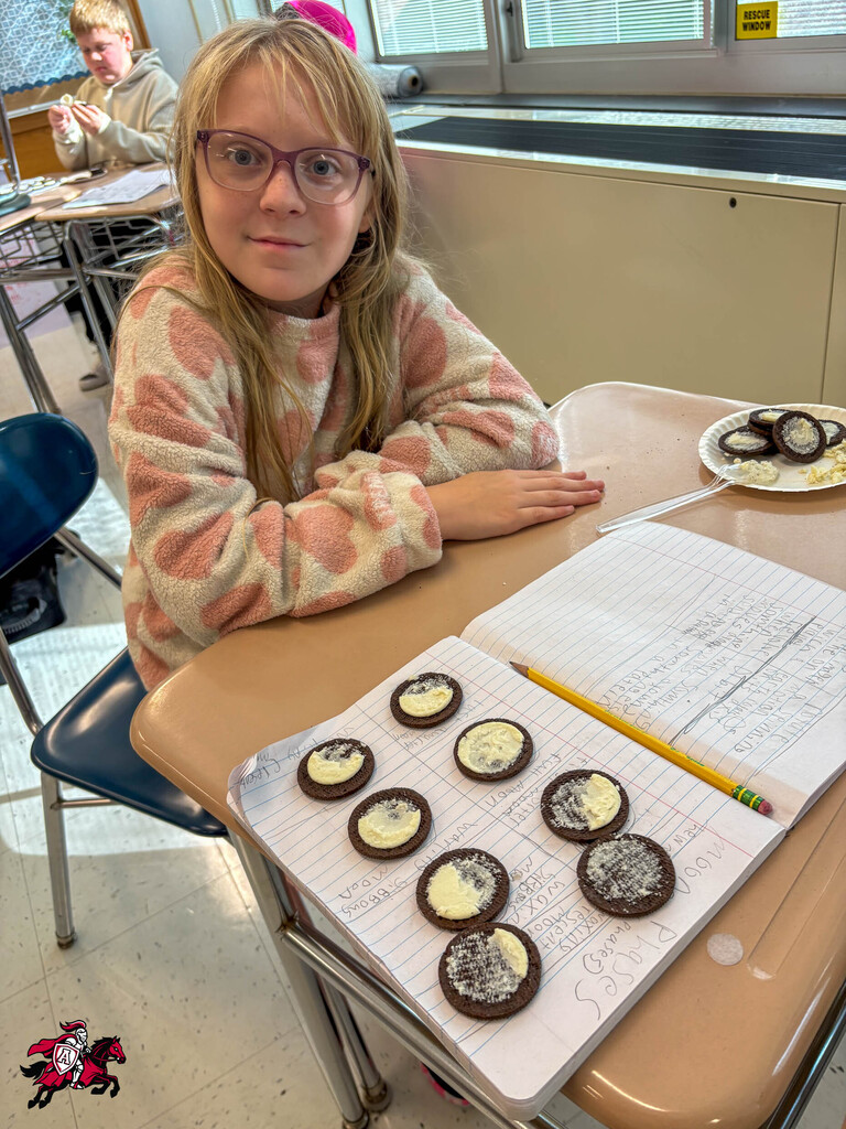 A girl sits at a table in a classroom. She has a pencil, a notebook, and cookies on the table.