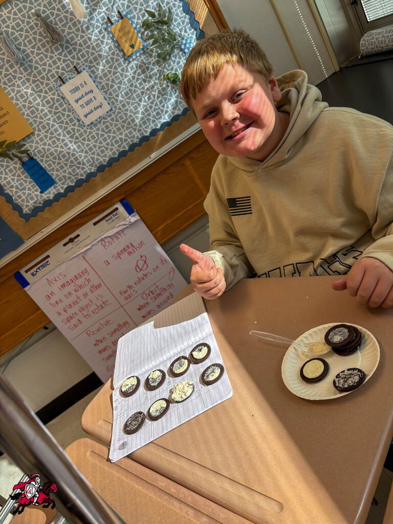A boy in a tan hoodie sits at a table, smiling and pointing. A plate of cookies sits on the table in front of him.