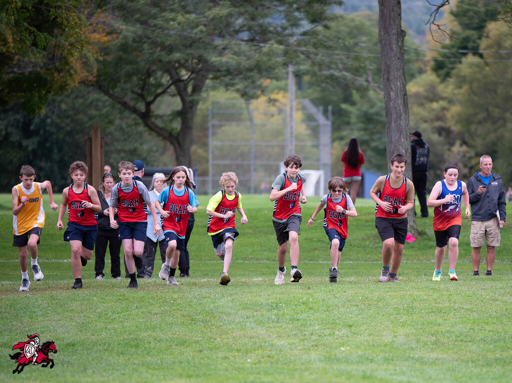 Children in red uniforms run across a grassy field with onlookers behind them, trees in the background.