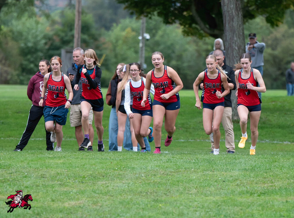 A group of young athletes in red and blue uniforms runs on a grassy field with trees and people watching.
