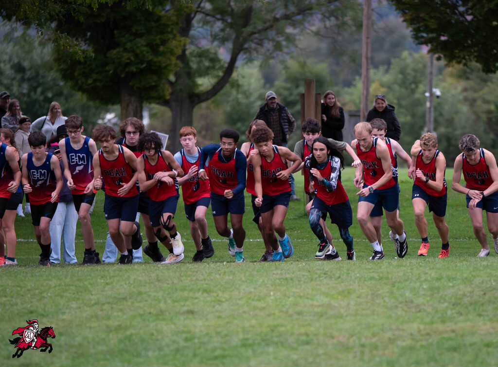 A group of runners in red and blue uniforms line up on a grassy field with trees in the background.
