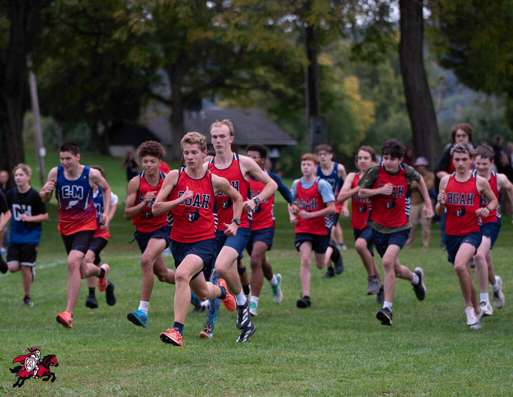 A group of runners in red and blue uniforms on a grassy field, with trees and spectators.