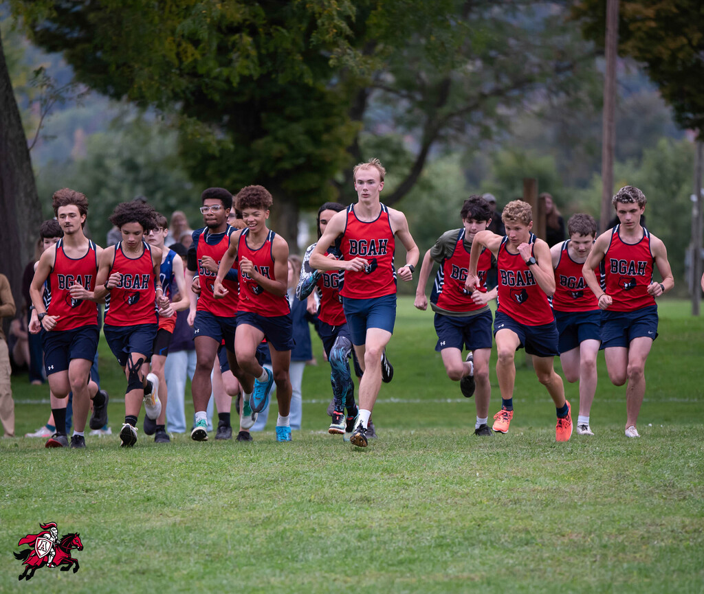 Cross country runners in red and blue uniforms run on a grassy field, with trees in the background.