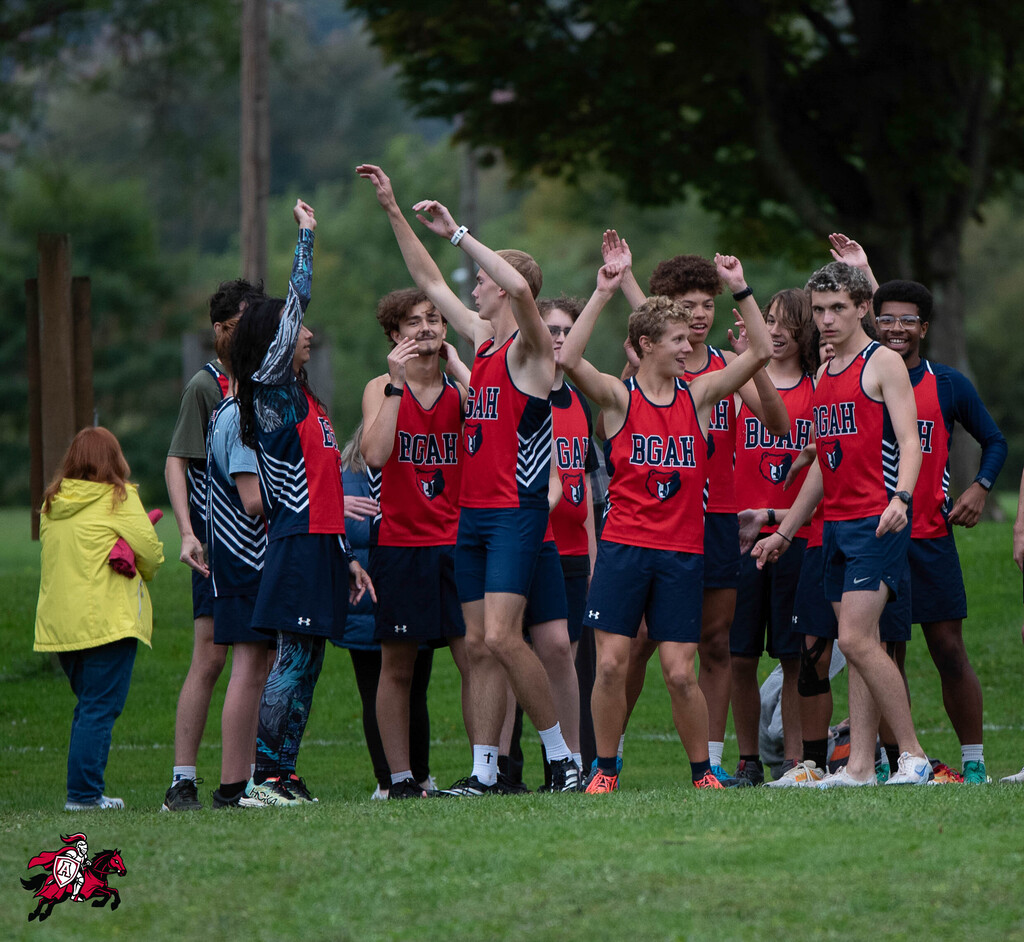 Team of athletes in red tops and blue shorts, some with raised hands, stand on a green field.