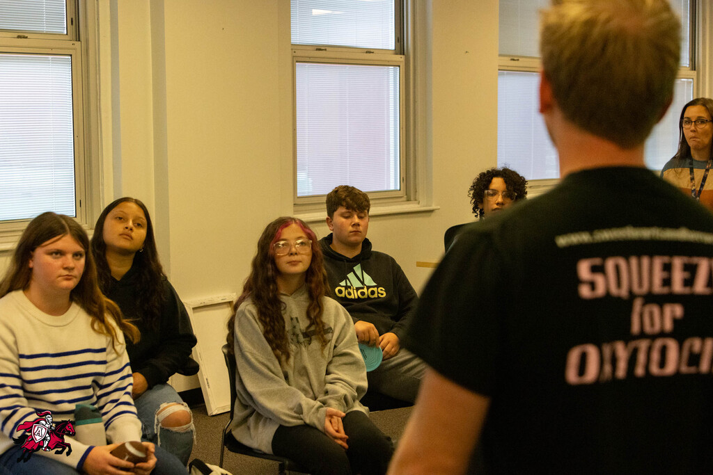 Five students sit in chairs, facing a person in a black shirt that says "SQUEEZE for OYTOCH."