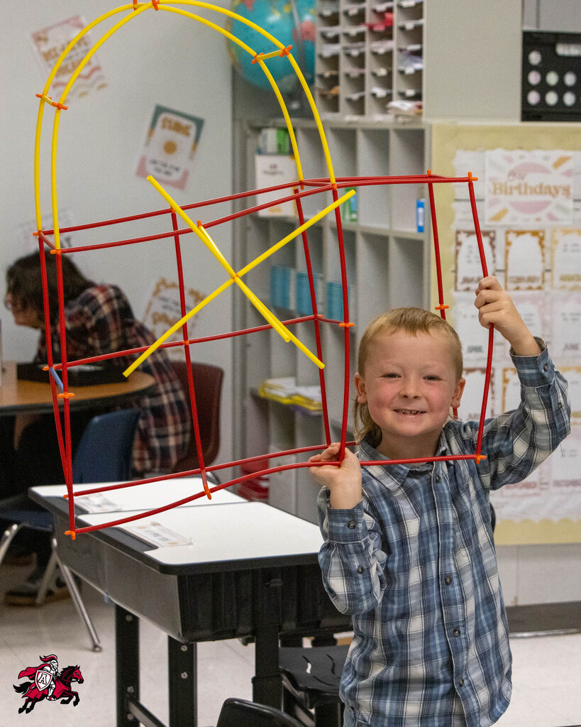 A child holds up a colorful geometric structure made of sticks, smiling. Background shows a classroom with desks and chairs.