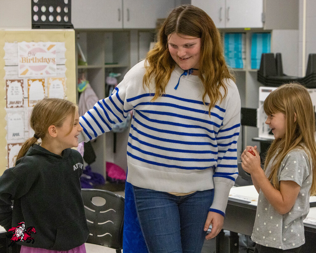 Woman with blue-white striped sweater and blue jeans holds two girls by shoulders. Background includes chairs and desks.
