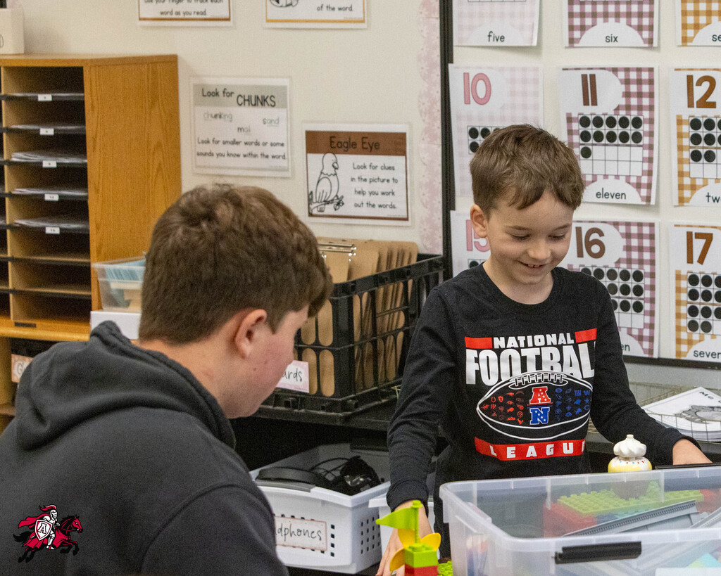 Two young boys in a classroom, one wearing a black shirt, the other with a football shirt, are engaged with a plastic container.