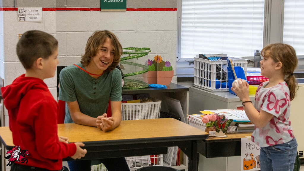 Three children seated around a table. A girl in a red hoodie and a boy in a green shirt.
