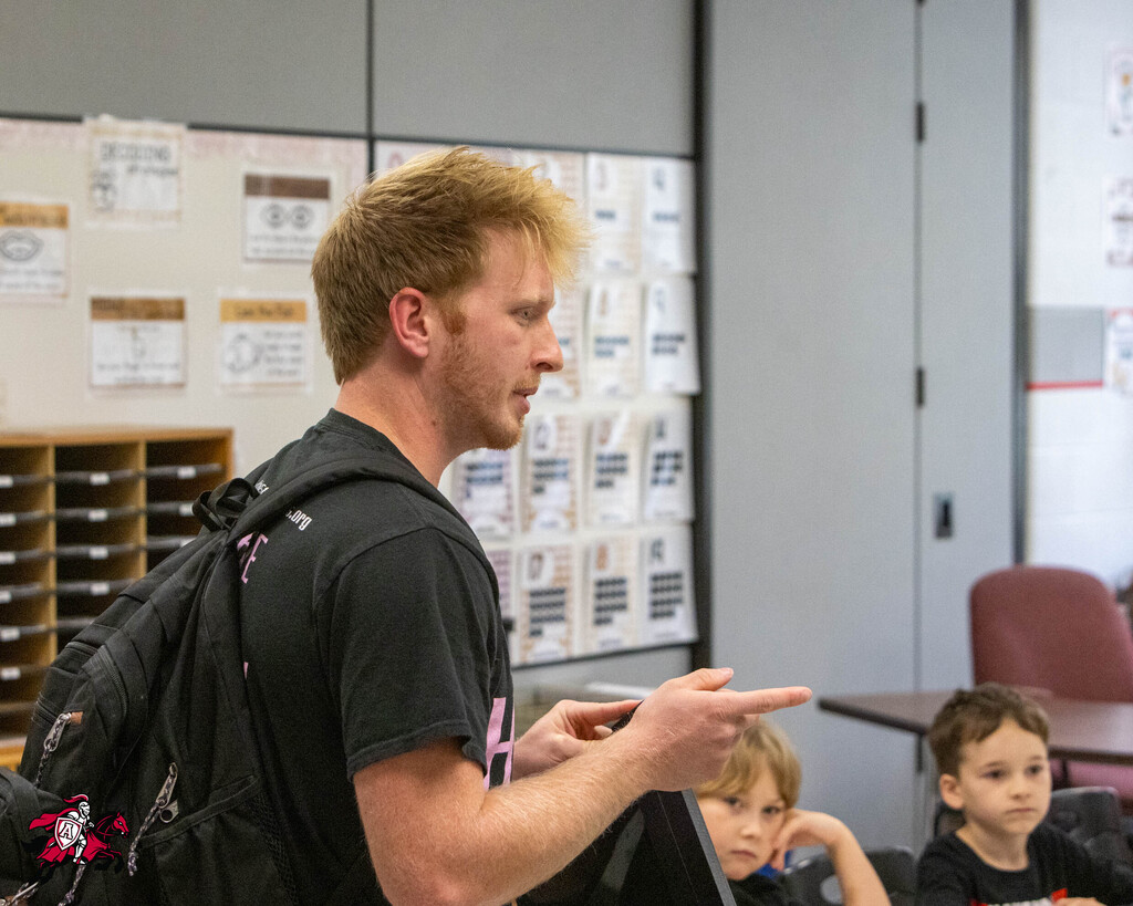 Man with backpack speaking to students. One student looks at him, the other looks down. Background includes bulletin board.