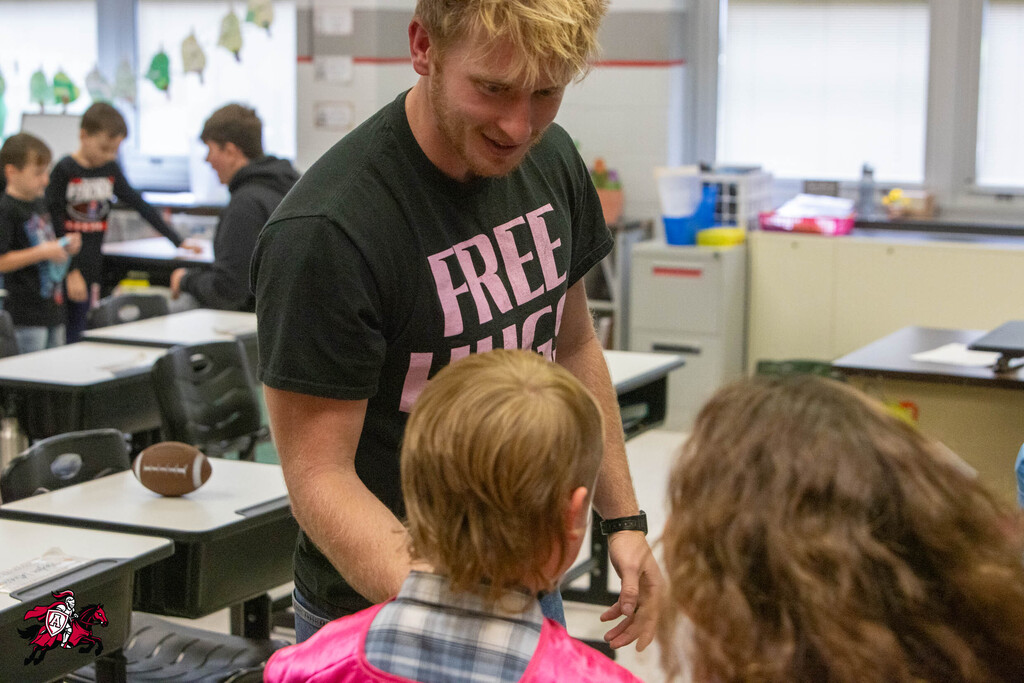 A person stands in a classroom with a child and another person, facing each other. Behind them, students sit at desks.