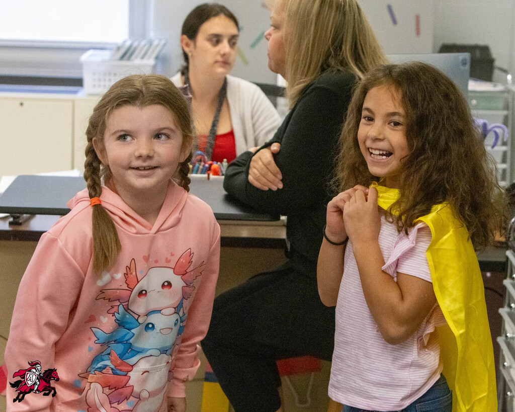 Two girls stand together, one with pigtails and a pink hoodie, the other with a yellow cape, smiling. A woman sits behind them, facing a desk.