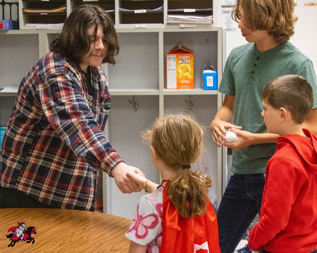 A classroom with three children and an adult. An adult in plaid holds a ball. The children look at the ball.