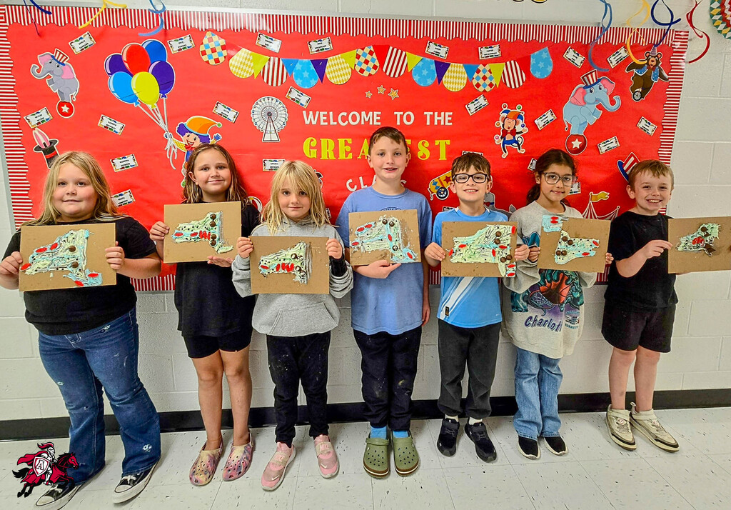 A group of children holds up painted maps in a classroom. Behind them, a banner says, "Welcome to the Great."